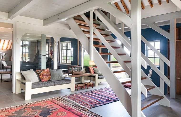 Open trap in Vakantiehuisje in Piaam leidt naar de bovenverdieping, met kleurrijke tapijten en comfortabele zitplaatsen.