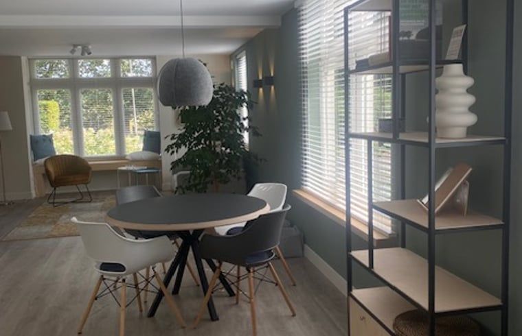 Essecke im Ferienhaus in Mander, Twente, Overijssel, mit einem runden Tisch und Blick auf den Garten durch grosse Fenster.